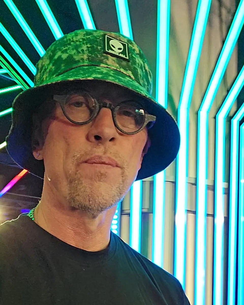 Norm Galarneau, who makes trance music as Norman Gordon, in a green bucket hat and round glasses, surrounded by teal neon light tubes