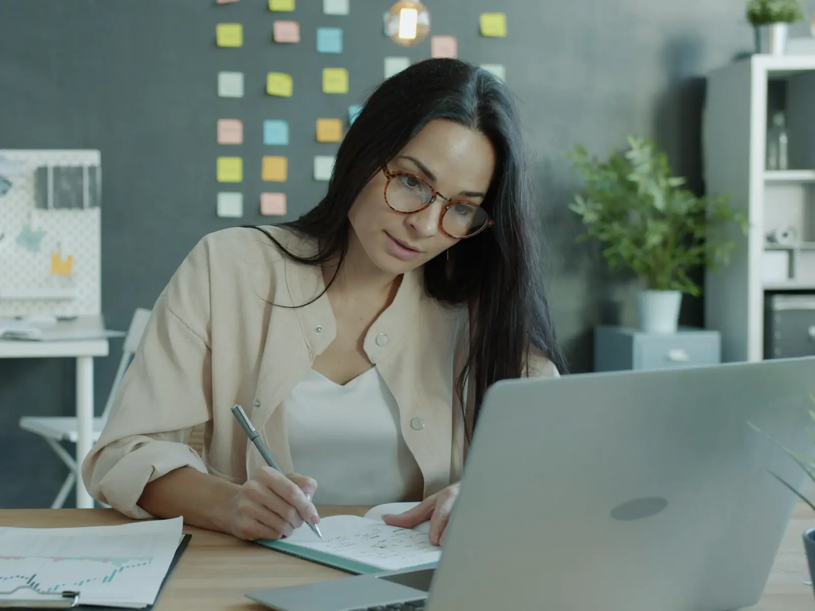 A woman taking notes while working on a laptop at her desk